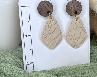May include: A pair of beige clay earrings with leaf and dot patterns, hanging from small, round, dark wood studs. The earrings are displayed on a white card with the text "Earring Designs By Sue". The earrings are approximately 1.5 inches long.