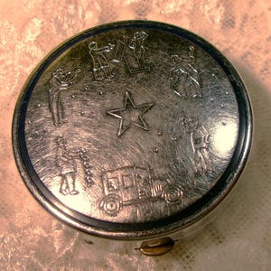 May include: A silver-toned compact with a blue rim. The compact features an engraved design of a star, a car, and several figures.