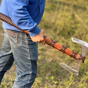Könnte beinhalten: Eine Person hält eine doppelseitige Axt mit einem Holzstiel, der mit braunem Leder umwickelt und mit Metallnieten befestigt ist. Die Axt hat einen rotbraunen Holzstiel und eine silberne Klinge.