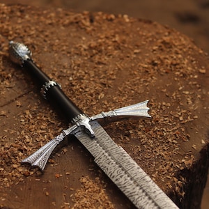 Peut inclure: Gros plan sur une épée avec une poignée noire et des détails argentés. La lame présente un motif texturé. L'épée repose sur une surface en bois parsemée de copeaux, créant une ambiance rustique.