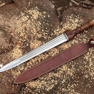 May include: A handcrafted dagger with a polished steel blade and a carved wooden handle. The dagger rests in a brown leather sheath, with wood shavings scattered around it. The handle features a detailed, woven design.