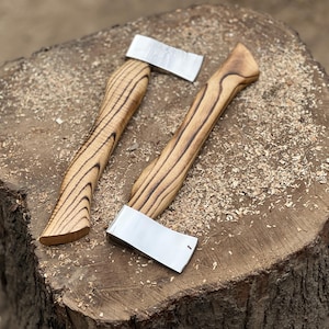 May include: Two small axes with wooden handles and silver blades rest on a tree stump covered in wood shavings. The handles have a light brown color with dark brown grain patterns. The blades are rectangular and shiny.