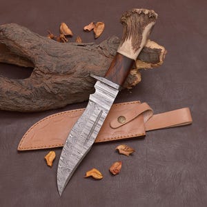 Puede incluir: Cuchillo de caza artesanal con una hoja de acero estampada y un mango de madera con empuñadura de asta. El cuchillo descansa sobre una funda de cuero, sobre un fondo marrón.