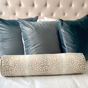May include: A beige and white patterned bolster pillow with a subtle ombre effect sits on a bed with blue velvet throw pillows.