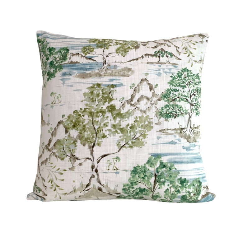 May include: A white cushion with a green and blue watercolour landscape design. The design features trees, mountains, and water.