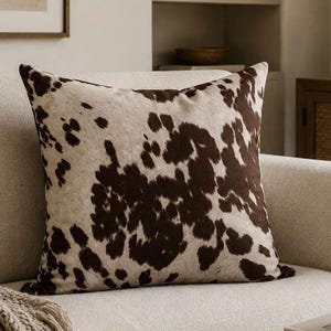 May include: A square throw pillow featuring a cowhide pattern with irregular dark brown patches on a light cream background. The pillow has a soft, textured appearance and is displayed in a standard upright position on a neutral-colored sofa.