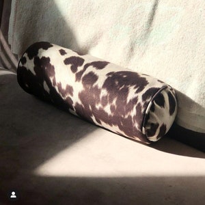 May include: A brown and white cowhide patterned cylindrical pillow. The pillow is on a light brown couch.