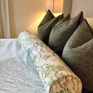 May include: A white and green patterned bolster pillow with a brown and green tree design. The pillow is on a white bed with a white and green patterned bedspread.