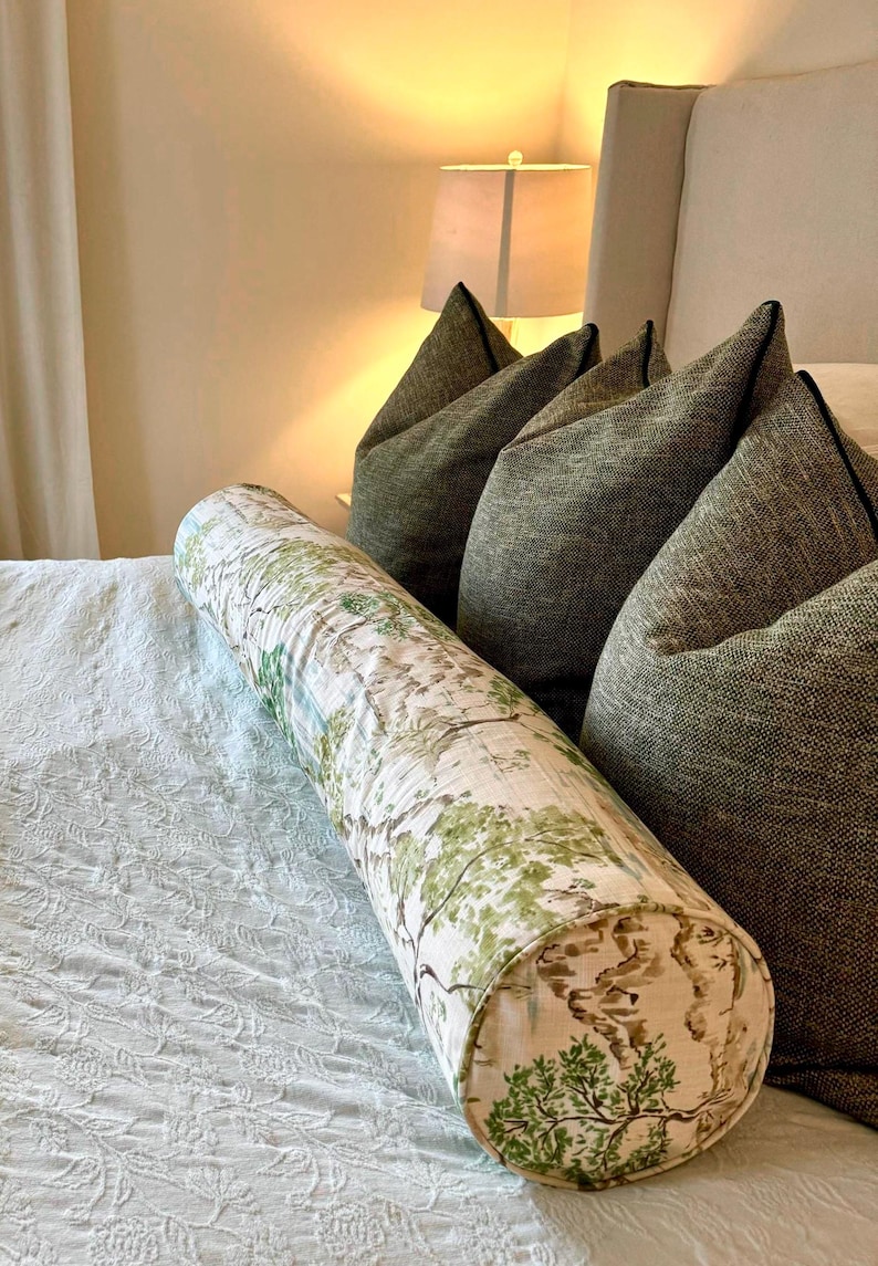 May include: A white and green patterned bolster pillow with a brown and green tree design. The pillow is on a white bed with a white and green patterned bedspread.