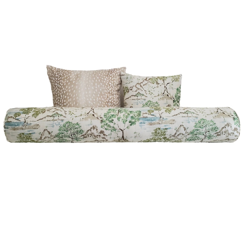May include: A cylindrical bolster cushion with a white background and a green and brown landscape print. Two decorative cushions are placed on top of the bolster cushion. One cushion is a light brown with a white spotted pattern, and the other cushion is white with a green and brown landscape print.