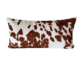May include: A long rectangular pillow with a brown and white cowhide print. The pillow is made of a soft, plush fabric.