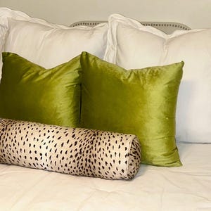May include: A bed with white pillows and two green velvet throw pillows. A cylindrical pillow with a black and tan animal print is in front of the green pillows. The bed is covered with a white sheet.