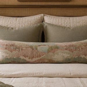 Decorative pastel landscape extra long lumbar pillow styled on a neutral bed with sage green accent pillows, woven textures, and warm traditional décor. This long linen lumbar pillow adds English cottage, countryside, grandmillennial, and heirloom-inspired charm to a bedroom while creating a soft, elegant, and inviting focal point.