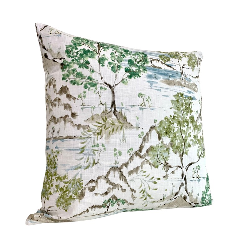 May include: A white cushion with a green and brown watercolour print of trees and mountains. The cushion is made of a soft, textured fabric.