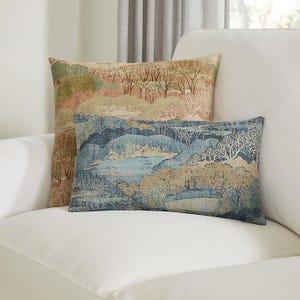 May include: Two decorative throw pillows with landscape designs. One pillow is square, the other is rectangular. The pillows feature a tapestry-like pattern with trees, hills, and a body of water in shades of blue, beige, and muted greens.