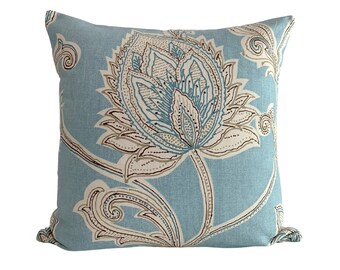 May include: A square throw pillow featuring a light blue fabric base with a large, intricate floral pattern in shades of white, dark blue, and brown. The design displays detailed botanical motifs with curved stems and petals across the surface.