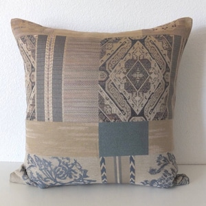 May include: A decorative throw pillow with a patchwork design featuring various shades of brown, beige, and blue. The pillow has a geometric pattern with intricate details.