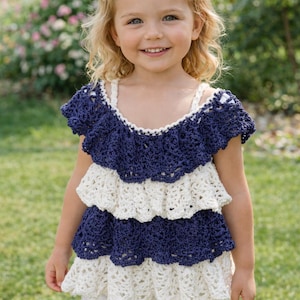 May include: A young girl wearing a handmade, tiered crochet top in navy blue and cream. The top has a ruffled design and is paired with white shorts. A white bow adorns her hair. The background is a green lawn and foliage.
