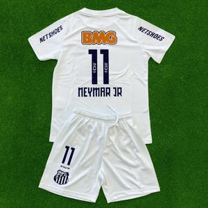 Pode incluir: Conjunto de camisa e calção de futebol brancos. A camisa apresenta o número 11 em azul marinho, o nome "NEYMAR JR" e os logotipos "BMG" e "NETSHOES". O calção também tem o número 11 e um logotipo da equipe.