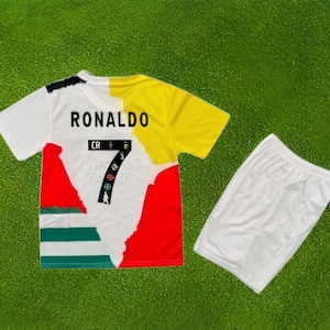 Puede incluir: Un conjunto de camiseta y pantalón corto de fútbol. La camiseta es blanca, amarilla, roja y verde, con el nombre "RONALDO" y el número "7". El pantalón corto es blanco liso. El conjunto está diseñado para el deporte.