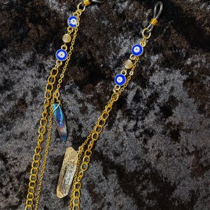 May include: Gold-coloured glasses chain featuring blue and white evil eye charms, small gold-coloured decorative elements, and two crystal pendants. The chain is made of multiple linked chains.