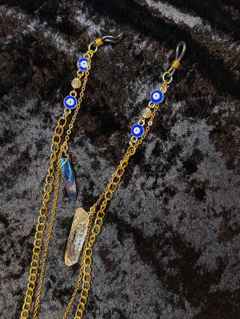 May include: Gold-coloured glasses chain featuring blue and white evil eye charms, small gold-coloured decorative elements, and two crystal pendants. The chain is made of multiple linked chains.