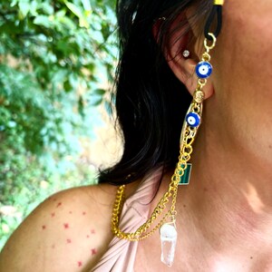 May include: A gold-coloured eyeglass chain with a black rubber loop. The chain features blue evil eye charms, a green square, and a clear crystal pendant. The chain is worn on the ear and drapes down the side.