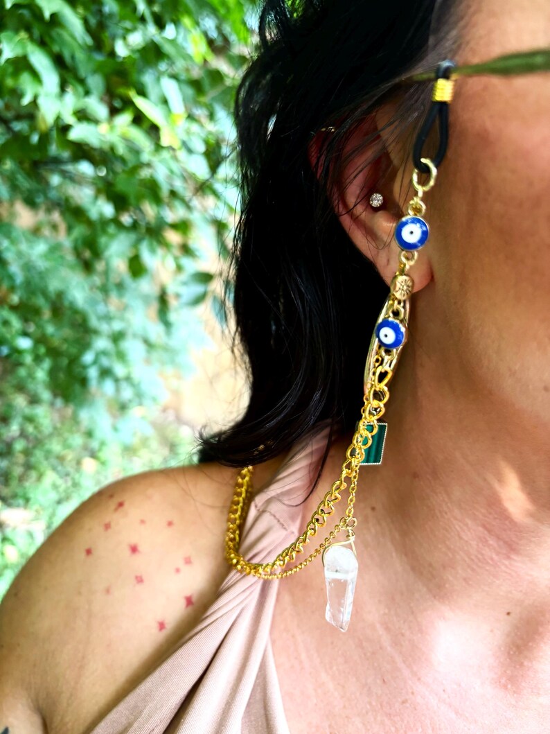 May include: A gold-coloured eyeglass chain with a black rubber loop. The chain features blue evil eye charms, a green square, and a clear crystal pendant. The chain is worn on the ear and drapes down the side.