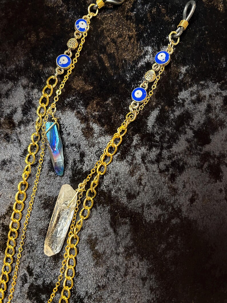 May include: Gold-coloured eyeglass chain with blue and white evil eye charms and two crystal pendants. The chain is made of multiple linked chains. One crystal is clear, and the other is iridescent blue and purple.