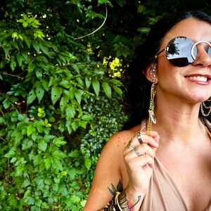May include: Circular sunglasses with a silver frame and dark lenses. The sunglasses have gold chains and crystal details attached. The person is wearing multiple rings and bracelets. Green foliage is in the background.