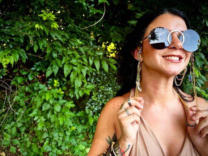 May include: Circular sunglasses with a silver frame and dark lenses. The sunglasses have gold chains and crystal details attached. The person is wearing multiple rings and bracelets. Green foliage is in the background.