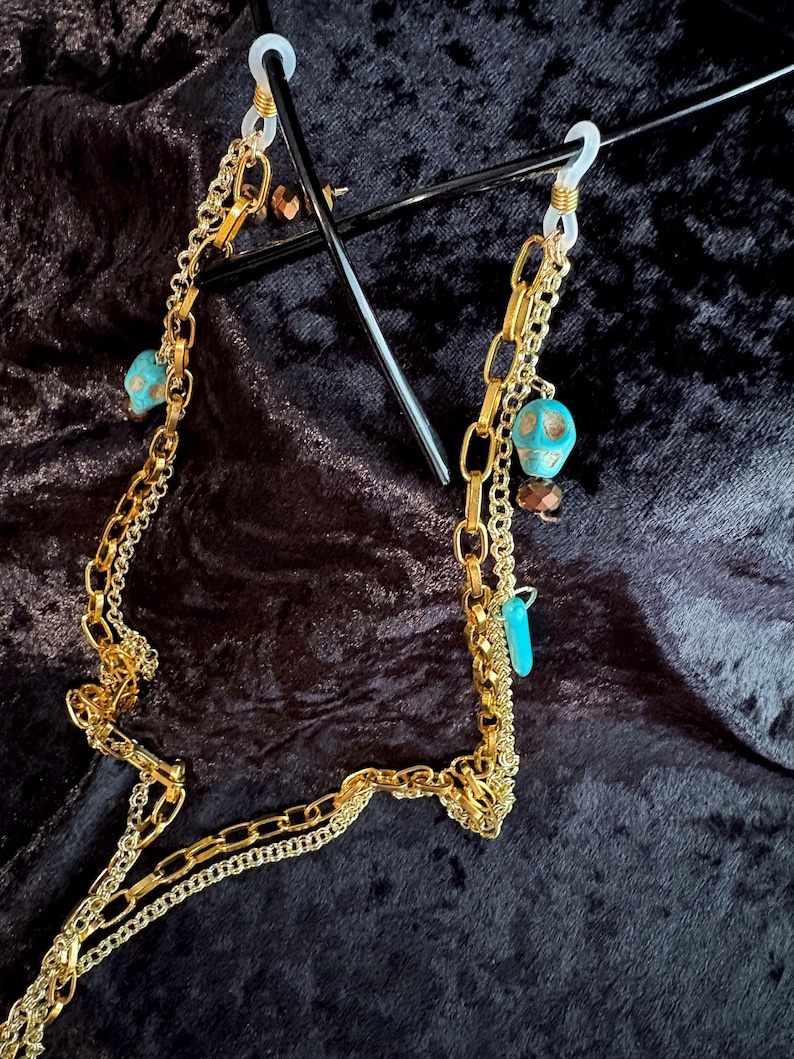 May include: Gold chain glasses holder with turquoise and skull beads. The chain is made of three different styles of gold chain. The chain is attached to clear plastic loops.
