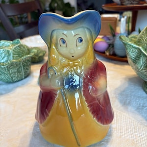 Puede incluir: Una figurita de cerámica de una mujer con un sombrero azul y un vestido rojo y amarillo. Lleva un palo en la mano derecha. La figurita está esmaltada en amarillo, rojo, azul y blanco.