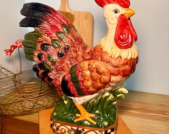 Pode incluir: Uma estatueta de galo de cerâmica com cores vibrantes. O galo tem uma crista e barbela vermelhas, um bico amarelo e um corpo multicolorido com penas vermelhas, verdes e castanhas. Ele está sobre uma base verde com uma borda decorativa. A estatueta está em uma superfície de madeira.