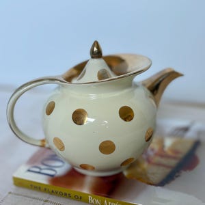 Puede incluir: Tetera de cerámica color crema con lunares dorados y detalles dorados. La tetera tiene un cuerpo redondeado, un asa curva y un pico. La tapa está rematada con un adorno dorado. La tetera está colocada sobre un libro titulado "The Flavors of Bon Appétit 2001".
