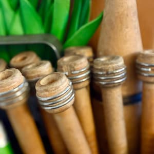 May include: A selection of antique wooden spools with metal rings, displayed in a wooden box. The spools are various shades of brown, with a cylindrical shape and a slightly flared top. Green leaves and pink flowers are visible in the background.