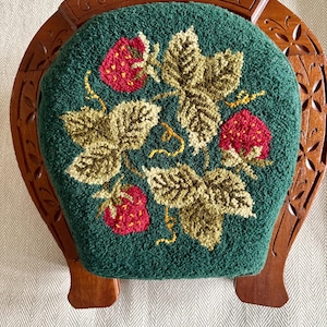 May include: A handcrafted wooden chair with a green cushion featuring a strawberry design. The cushion has a textured appearance with red strawberries and green and tan leaves. The chair's back is arched and has the words "GOOD LUCK" carved into it.