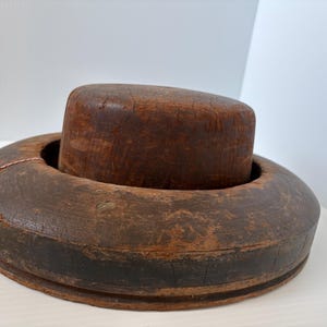 May include: A vintage hat block, featuring a dark brown, cylindrical form set within a circular base. The wood shows visible grain and signs of age, with a tag attached. This antique tool is likely used for shaping hats.