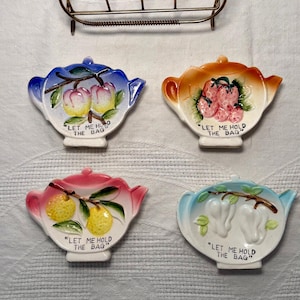 Puede incluir: Un juego de cuatro posa-bolsas de té de cerámica con forma de tetera, cada uno con un diseño de fruta diferente y el texto "LET ME HOLD THE BAG". Un portabolsas de té de metal está encima de las piezas de cerámica.