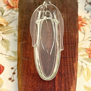 May include: A clear glass dish shaped like an ear of corn with a textured husk design on the exterior. The dish has an elongated oval form with a pointed top and a rounded base, featuring molded details that mimic the kernels and leaves of corn.