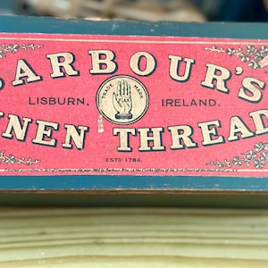 May include: A rectangular box with a dark teal base and a red label featuring the text BARBOUR'S LINEN THREAD, LISBURN, IRELAND, ESTD 1784. The label includes a central hand icon with the word FLAX and various circular award seals.