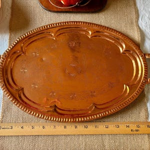 Può includere: Vassoio ovale color rame con manici. Il vassoio presenta un motivo floreale decorativo e un bordo perlato. Un righello di legno è posto sotto, che misura fino a 45 cm.