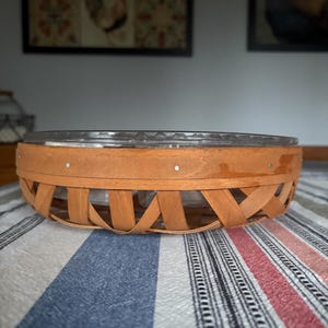 May include: A woven basket with a clear glass insert. The basket is made of natural wood and has a brown stain. The basket is sitting on a striped fabric.