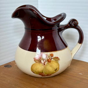 Puede incluir: Una jarra de cerámica vintage con un diseño de dos tonos. La parte superior es de color marrón oscuro brillante, mientras que la parte inferior es de color crema con una ilustración de fruta. El asa es de color crema con un acento marrón oscuro. La jarra tiene un pico único y ondulado.