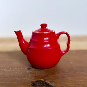 May include: A small, glossy red teapot with a curved spout, a rounded body, and a loop handle. The lid has a small knob. The teapot is sitting on a wooden surface.