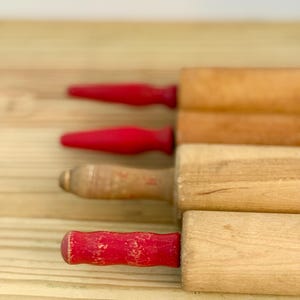 Può includere: Una collezione di quattro mattarelli in legno disposti orizzontalmente. Due mattarelli presentano manici lisci rosso brillante, uno ha un manico in legno naturale usurato con dettagli in metallo e quello in basso ha un manico rosso scheggiato.
