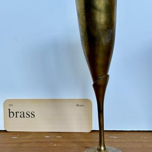 Puede incluir: Una flauta de champán de latón vintage con un tallo delgado y una parte superior ancha y acampanada. La flauta tiene un diseño retorcido único en el medio. Una tarjeta con la palabra "brass" está al lado de la flauta.