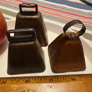 May include: Three antique cowbells, one with a copper finish and two with a dark brown finish. The cowbells are sitting on a white surface with a wooden ruler in the foreground.