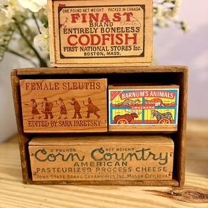 May include: A wooden shelf unit containing three vintage-style wooden boxes. The top box reads FINAST BRAND ENTIRELY BONELESS CODFISH. The middle boxes feature FEMALE SLEUTHS and BARNUM'S ANIMALS. The bottom box reads CORN COUNTRY AMERICAN CHEESE.