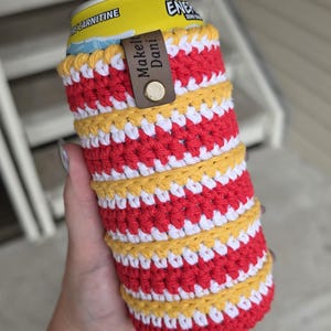 Può includere: Un portabicchieri all'uncinetto a righe rosse, bianche e gialle. Il portabicchieri ha un'etichetta in pelle marrone con la scritta "Makell Dani" e un bottone automatico in metallo. Progettato per una lattina di bevanda di dimensioni standard.